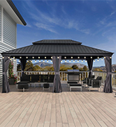 avec toit en métal noir sur terrasse en bois. Structure de support à piliers gris. Lampes suspendues visibles à l'intérieur. Fond de ciel bleu avec bord de maison blanc visible.