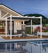 Terrasse extérieure avec pergola blanche, adjacente à une piscine. Les sièges comprennent des chaises en osier et une table. L'extérieur de la maison est visible avec des accents de pierre.